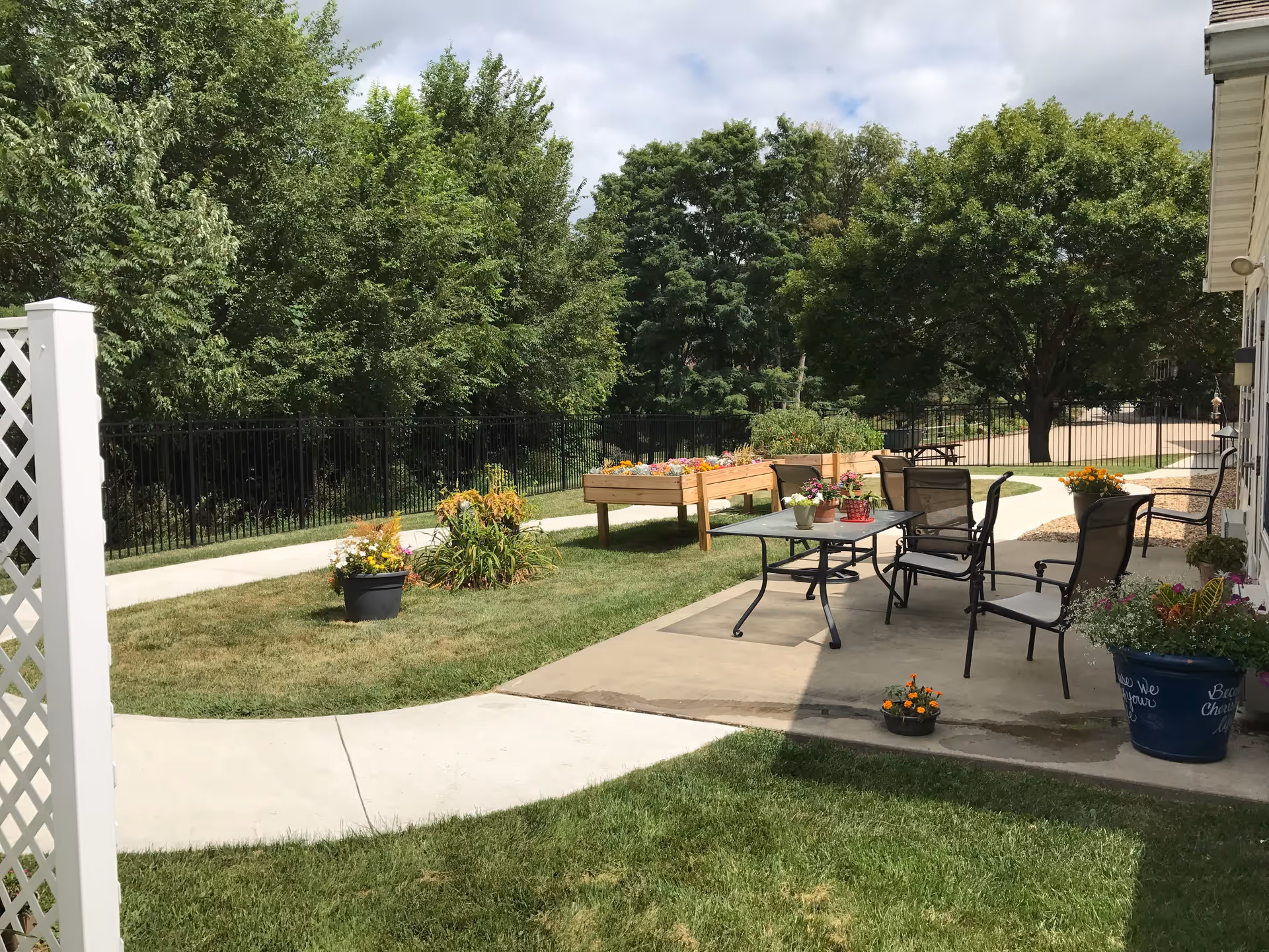 Outdoor patio area with a concrete walkway, several chairs and a glass-top table with potted flowers. Raised garden beds with colorful flowers and various potted plants are visible. The area is surrounded by green grass, trees, and a black metal fence under a partly cloudy sky.