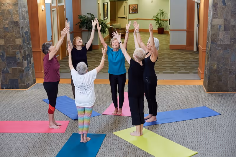Six elderly women standing barefoot on colorful yoga mats in a circle indoors, raising their arms upwards as part of a group exercise or yoga session in a senior living facility.