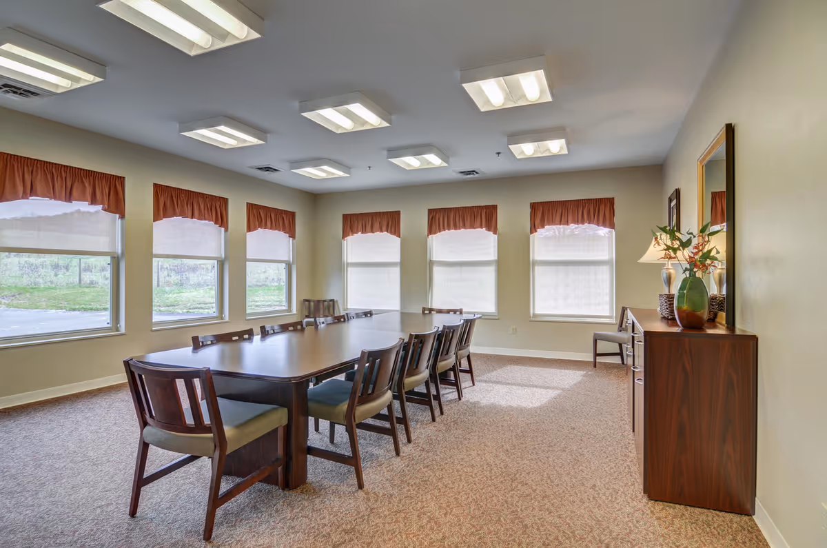 A bright dining/meeting room with a long rectangular table surrounded by chairs, several windows with valances, and a sideboard with decorative vases and a mirror.