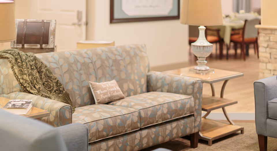 A cozy living room area with a patterned sofa, a decorative throw blanket, and a small pillow with text. There is a side table with a lamp next to the sofa, and in the background, a dining table with chairs is visible.
