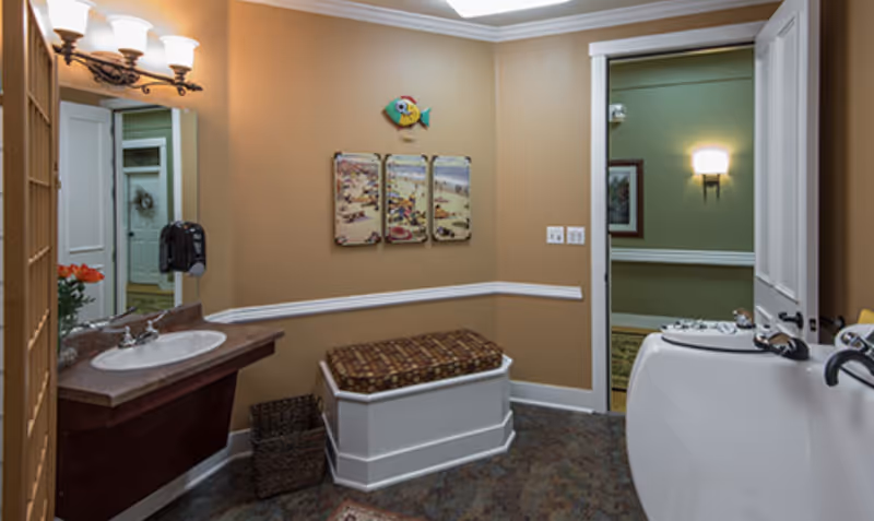 A bathroom in an assisted living facility featuring a sink with a mirror and light fixture above it on the left, a cushioned bench with a patterned seat against a beige wall, a decorative fish wall hanging, and a white bathtub with handrails on the right. The room has a brownish floor and white trim, with an open door leading to a green-painted adjacent room.