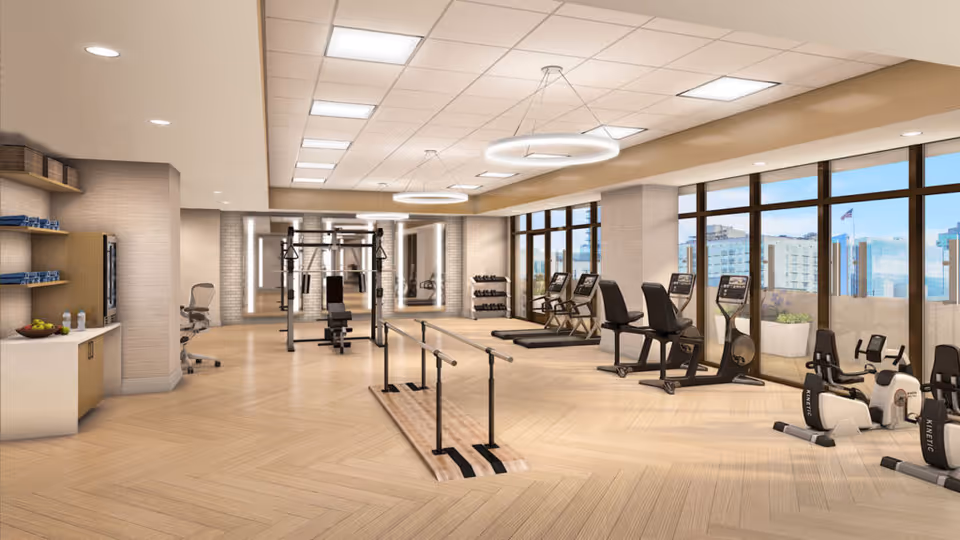 A bright, modern fitness room with cardio machines, weight equipment, a barre, and large windows overlooking the city.