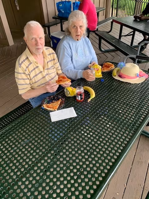 Two elderly people seated at a covered outdoor picnic table eating pizza, with drinks, a banana and a sun hat on the table.
