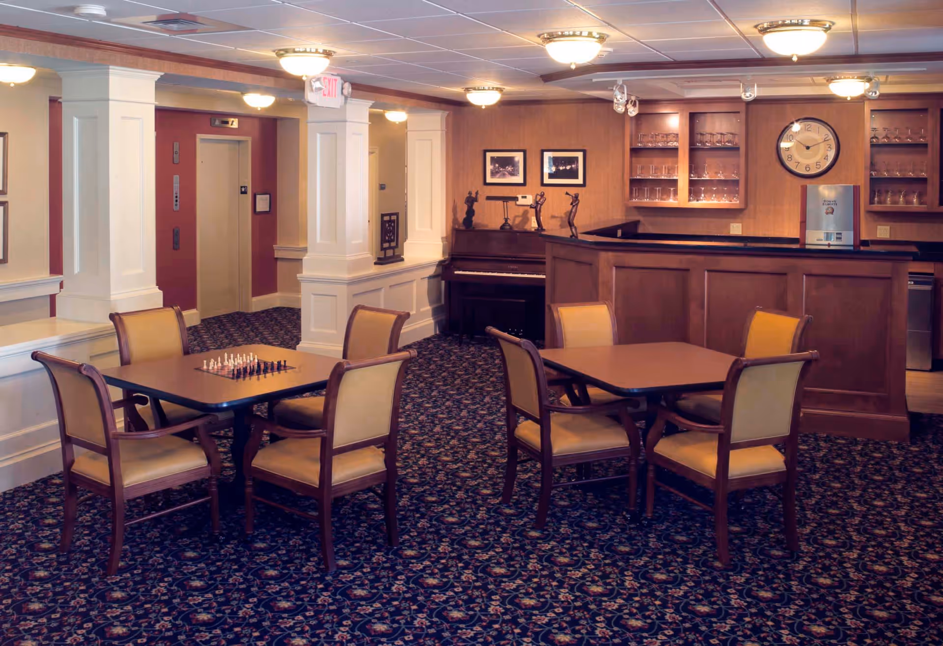 Interior of a senior living facility common area with two tables each surrounded by four chairs. One table has a chessboard set up. There is a carpet with a floral pattern, a wooden bar with glass shelves holding glassware, a clock on the wall, and a piano in the corner. Two elevators are visible in the background.