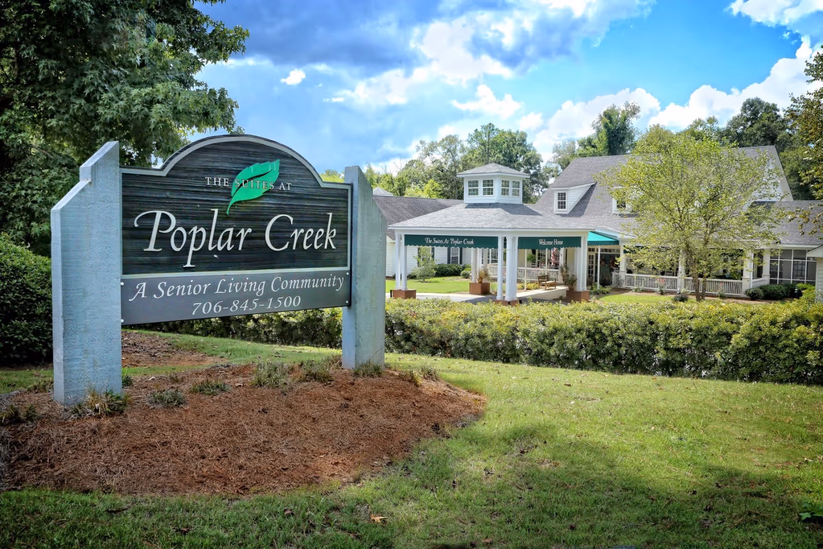 Outdoor view of Poplar Creek Senior Living Community entrance sign with a large green leaf logo, phone number, and a white building with a covered porch and green awnings in the background surrounded by trees and bushes under a partly cloudy sky.