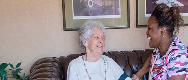 An elderly woman with white hair sitting on a brown leather couch smiling while a healthcare worker in a colorful scrub top checks her blood pressure with a cuff and stethoscope in a cozy room with framed pictures on the wall.