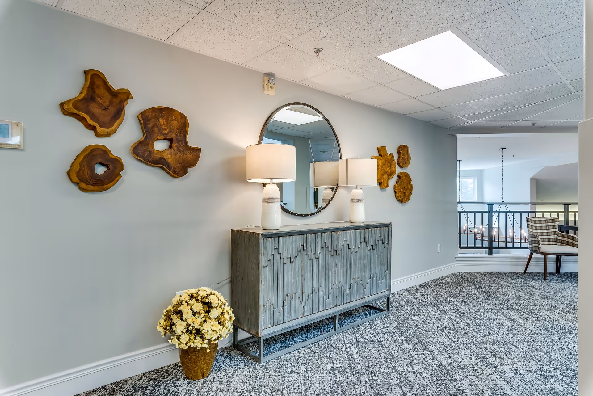 A hallway area in a senior living facility featuring a decorative wooden sideboard with two white lamps on top, a round mirror mounted on the wall above it, and three wooden wall art pieces. A pot of yellow flowers is placed on the floor next to the sideboard. The space has carpeted flooring, light gray walls, and a ceiling with recessed lighting. In the background, there is a railing and a chair with a plaid cushion near a window.