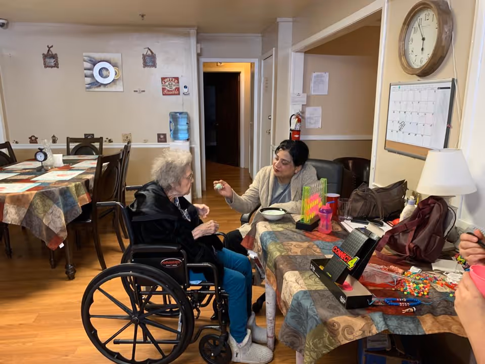 An elderly woman in a wheelchair is interacting with a caregiver seated at a table covered with a patterned tablecloth. The table has a Connect 4 game, a pink sippy cup, and various small items. In the background, there are more tables and chairs, a water dispenser, wall decorations, a clock, and a calendar on the wall.