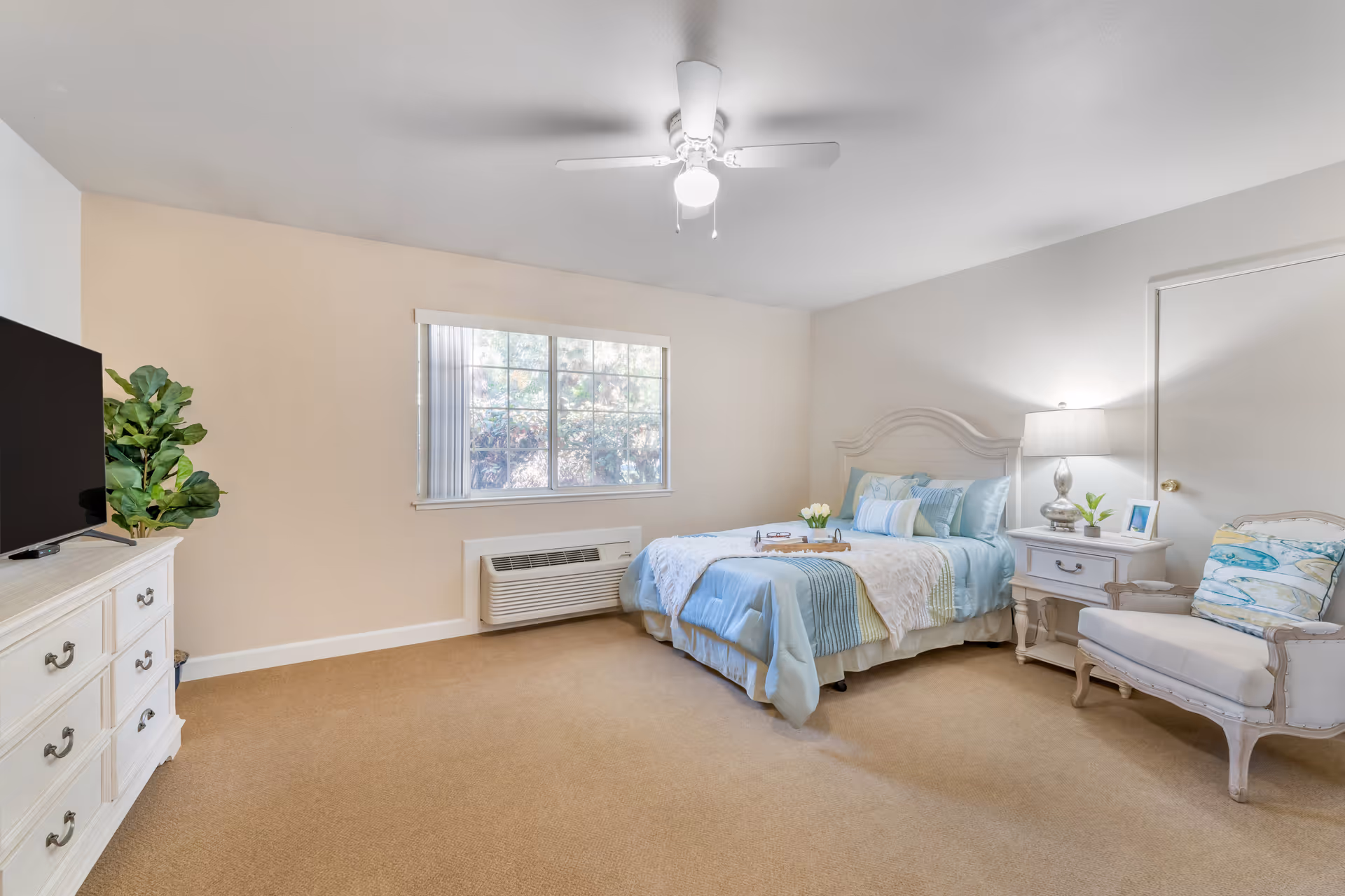 A bright and cozy bedroom with a bed dressed in blue and white linens, a white nightstand with a lamp and small plant, a cushioned armchair with a decorative pillow, a white dresser with a TV on top, a large window with vertical blinds, and a ceiling fan with a light.