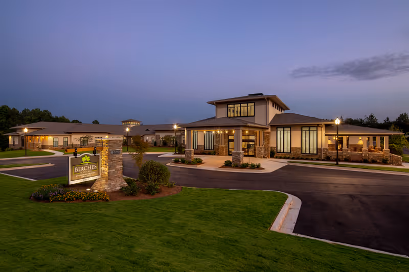 Front exterior of The Birches at Villa Rica senior living facility at dusk, showing the illuminated entrance, sign, and landscaped lawn.