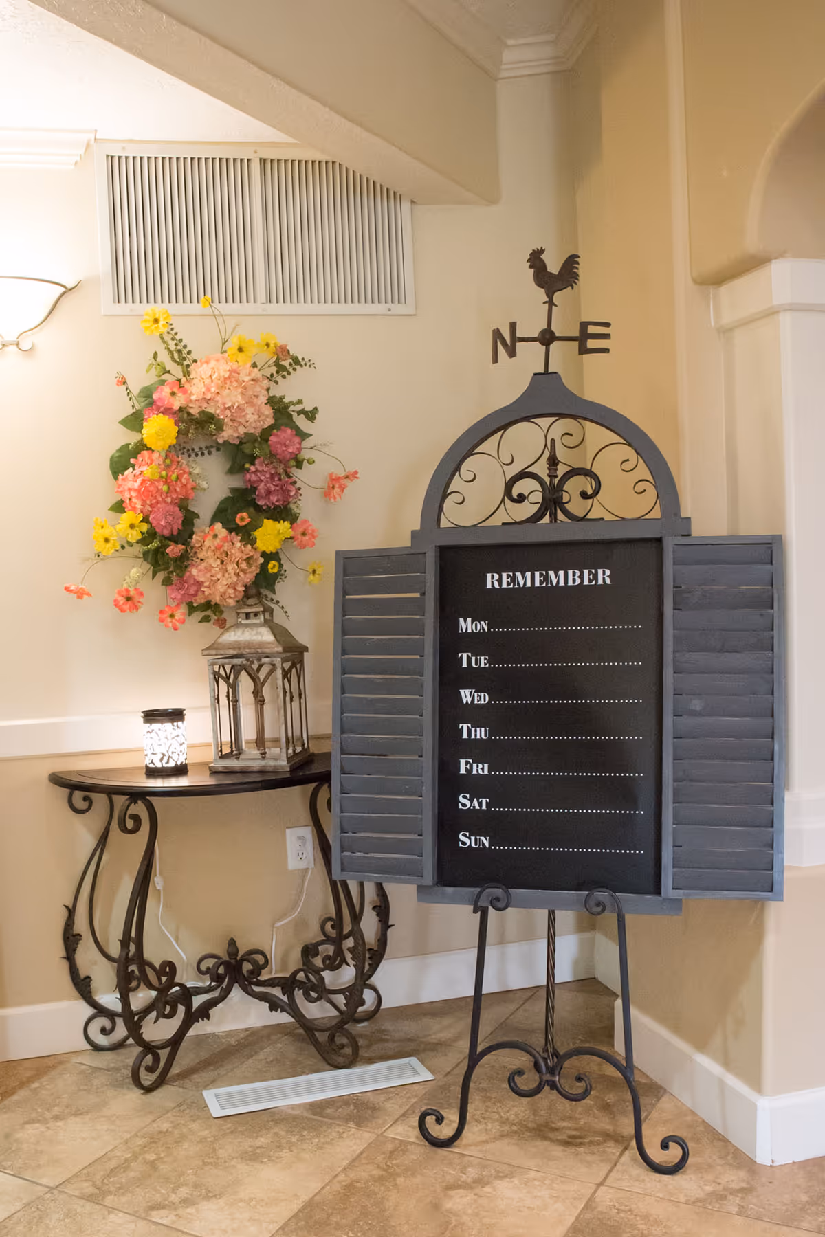 An interior corner of a room featuring a decorative black metal stand with a blackboard labeled 'REMEMBER' and days of the week listed below. To the left, there is a small ornate table with a colorful floral wreath and a lantern on top. The floor is tiled and the walls are painted a light beige color.