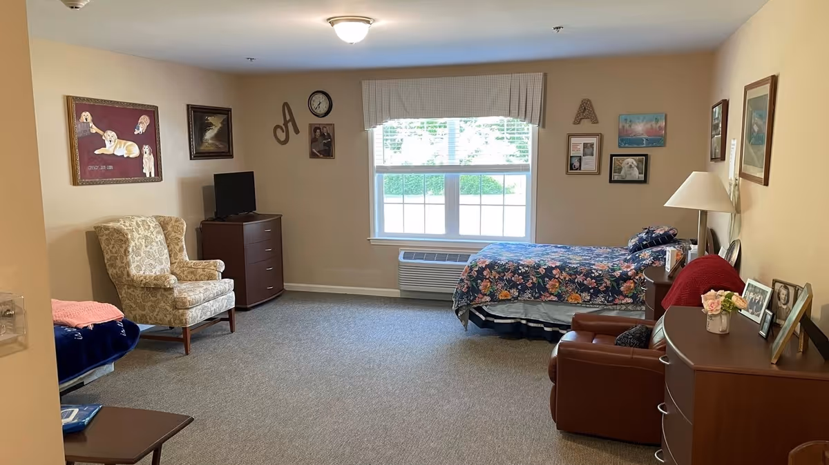 A cozy bedroom in a senior living facility with a single bed covered in a floral bedspread near a window with blinds and a valance. The room has beige walls decorated with framed pictures and wall art, a floral upholstered armchair, a small TV on a wooden dresser, a leather recliner, and another wooden dresser with family photos and a lamp.