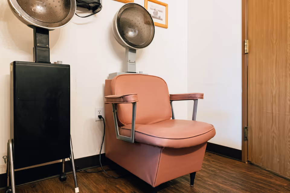 A vintage pink salon chair with armrests positioned next to two black and silver hair drying machines mounted on stands, set against a white wall with a wooden door on the right and a framed picture hanging above.