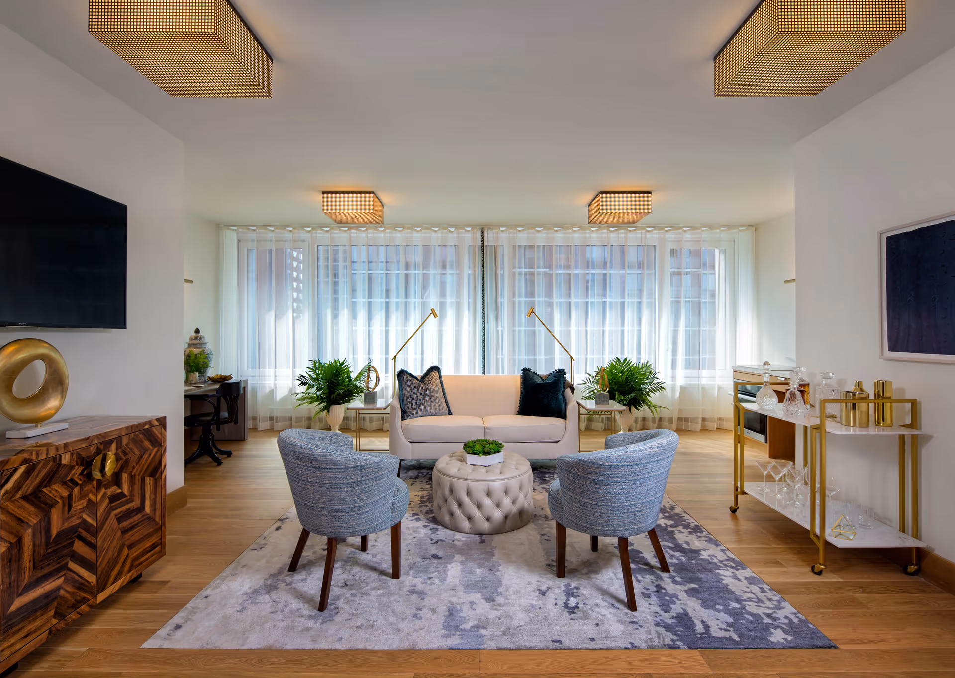 Modern, well-lit sitting room with a sofa, two upholstered armchairs around a tufted ottoman, decorative plants, and large sheer-covered windows.