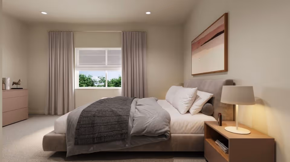 A neatly made bed with gray bedding and white pillows in a softly lit bedroom. There is a wooden nightstand with a lamp and some items on it to the right of the bed. A large window with curtains is centered on the back wall, showing greenery outside. A dresser with decorative items is on the left side of the room, and a framed abstract painting hangs above the bed.