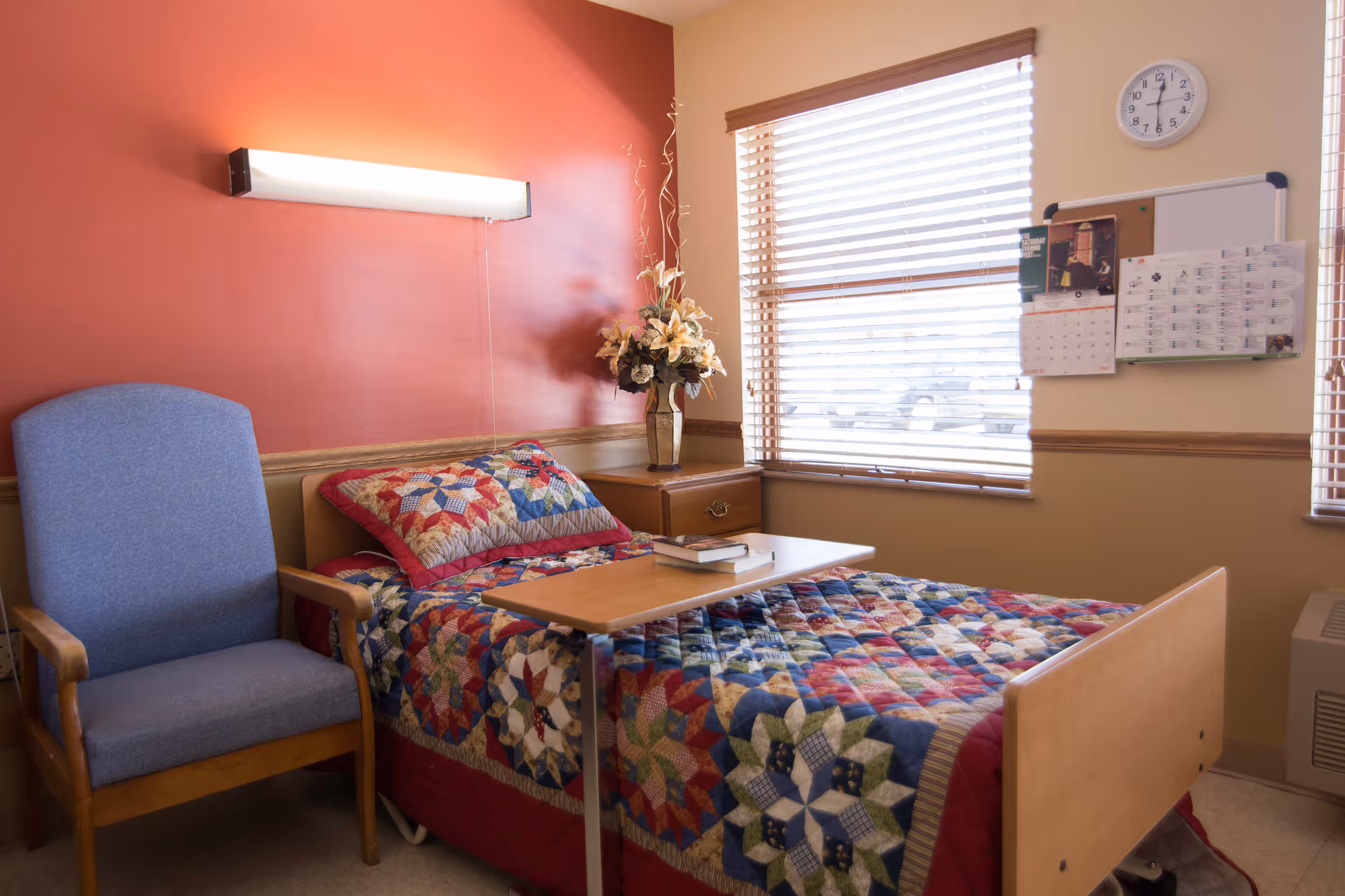 A small, cozy room with a single bed covered in a colorful patchwork quilt. Next to the bed is a wooden nightstand with a vase of flowers and a blue upholstered armchair. A wooden tray table is positioned over the bed with two books on it. The room has a window with blinds, a wall clock, and a bulletin board with a calendar and papers pinned to it. The walls are painted in warm tones, with one wall in a reddish color and the others in beige.