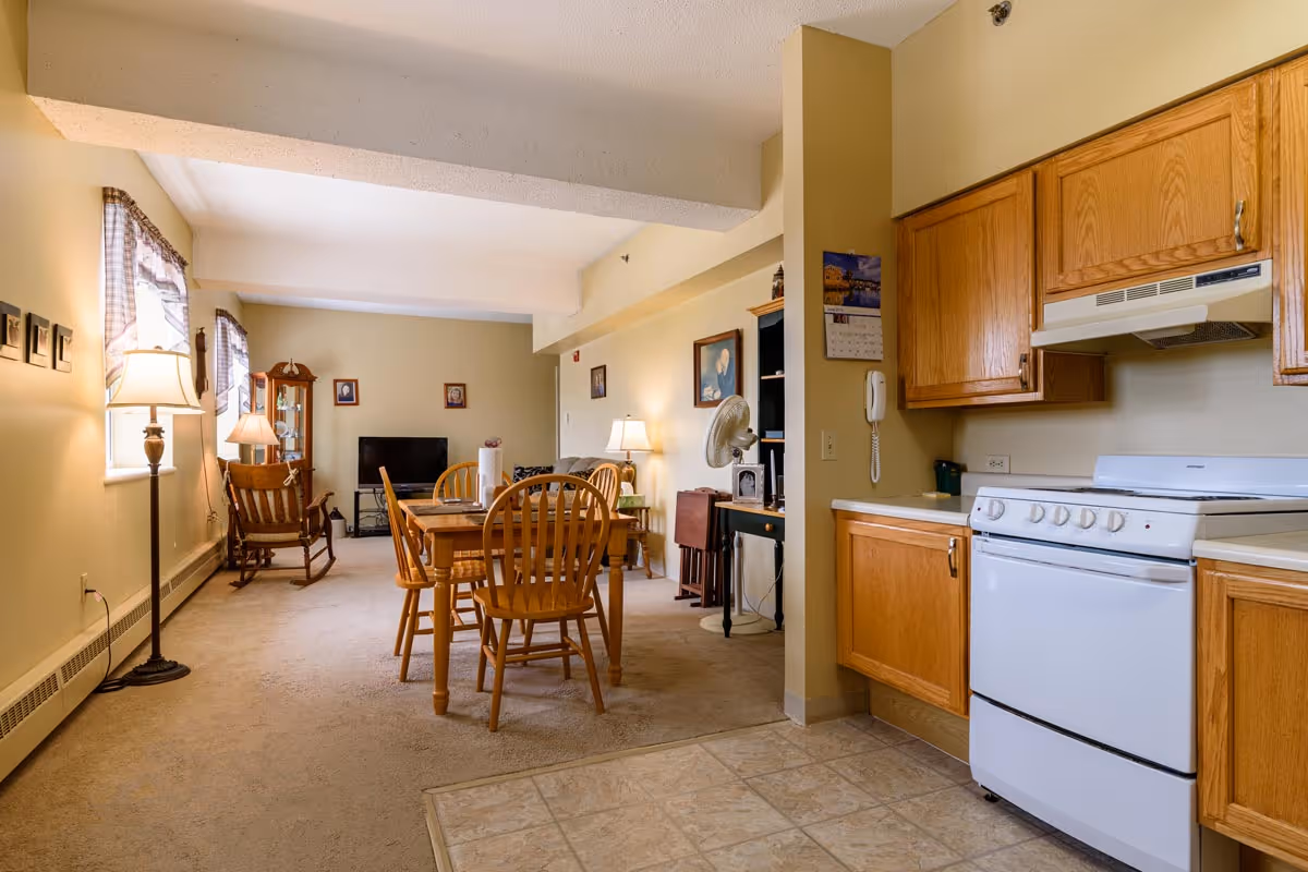 Open interior showing a small kitchen with oak cabinets and a white stove opening into a dining area with a wooden table and chairs and a living area with a TV and rocking chair.