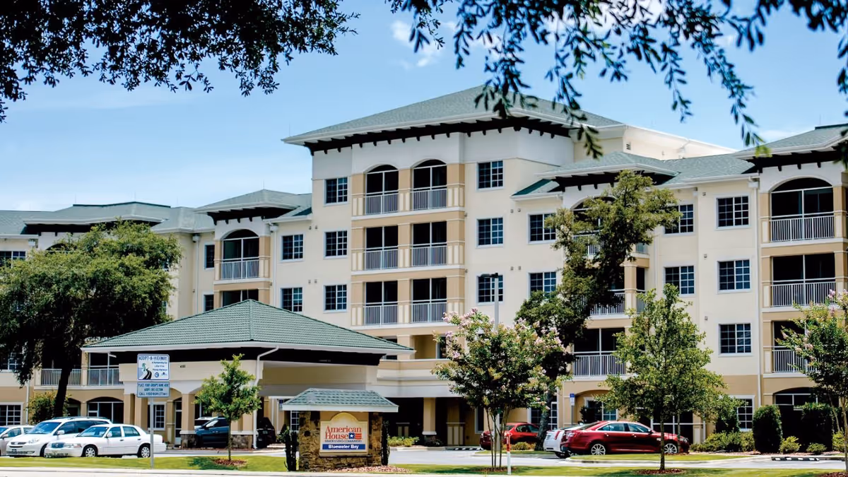 Exterior view of a multi-story senior living facility named American House Bluewater Bay with a green roof, balconies, trees, and parked cars in front under a blue sky.