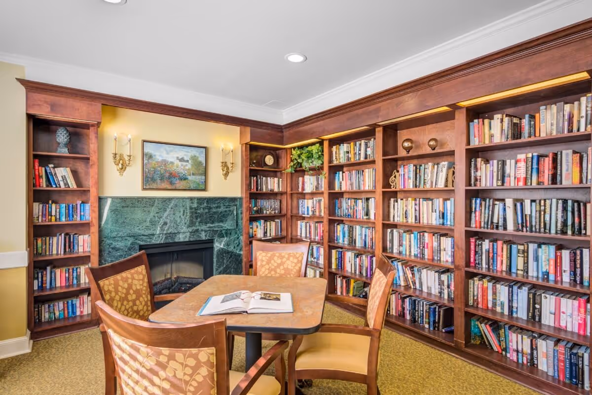 Cozy library/reading room with built-in wooden bookshelves, a marble fireplace, and a table surrounded by chairs.