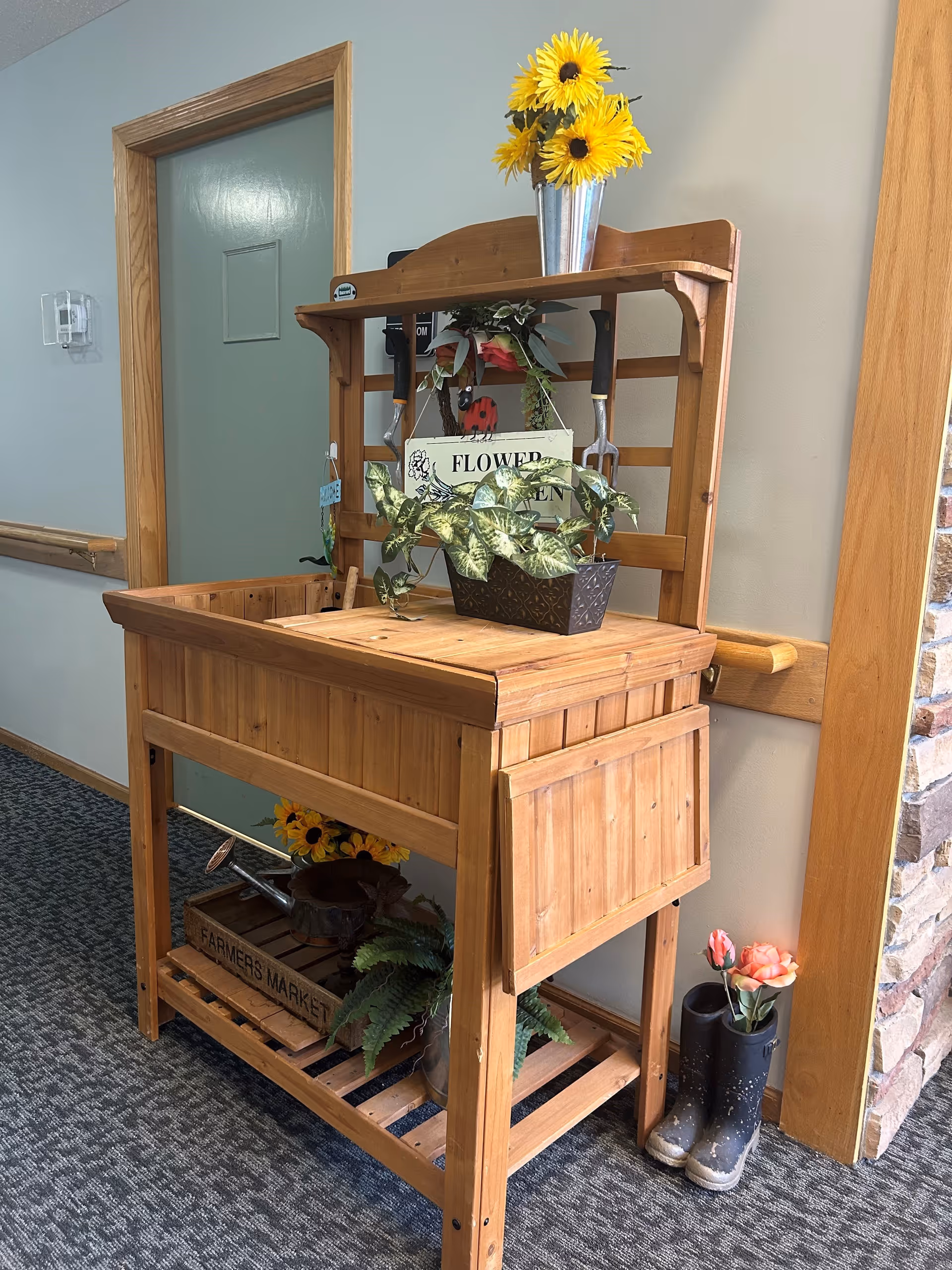 A wooden gardening potting bench with gardening tools hanging on the back panel, a metal bucket with yellow sunflowers on top, and various potted plants and flowers arranged on and under the bench. A pair of black rain boots with pink flowers inside is placed on the floor next to the bench. The bench is situated in a hallway with a green door and light-colored walls.