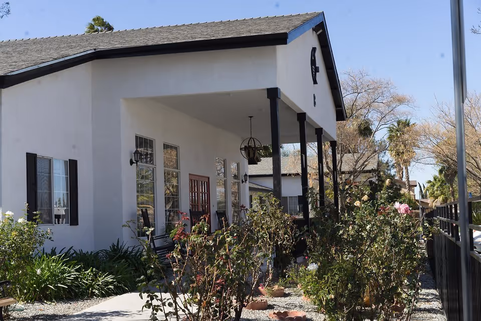 Exterior view of a white building with a covered porch supported by black pillars. The porch has hanging light fixtures and several windows with black shutters. There are various plants and bushes in front of the building, and a black metal fence runs along the right side. Trees and a clear blue sky are visible in the background.