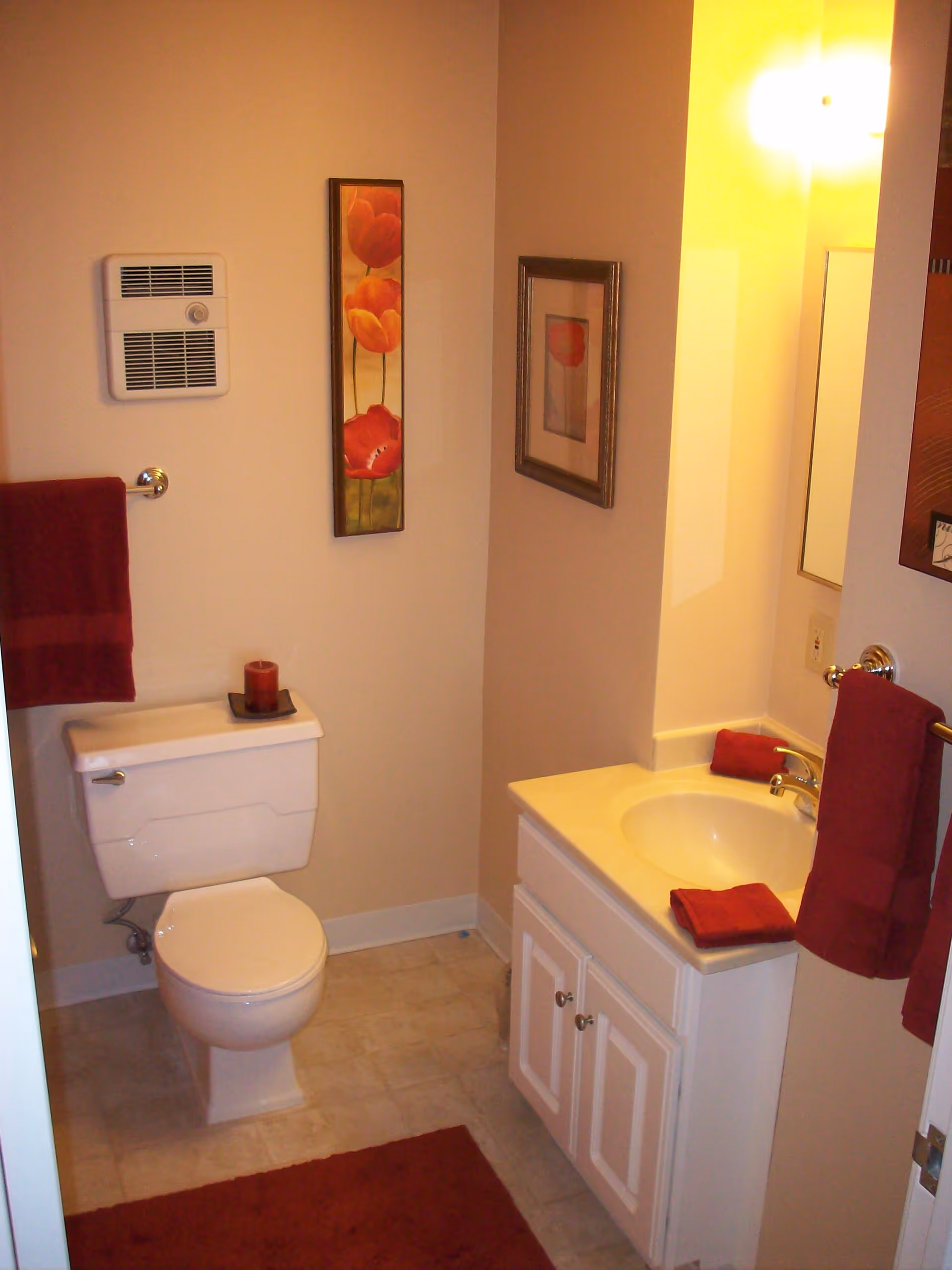 A small bathroom with a toilet, white vanity sink and mirror, red towels and decorative wall art.