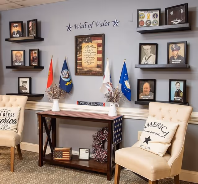 A cozy room corner with two beige upholstered chairs, each with a patriotic pillow. Between the chairs is a wooden console table decorated with small American flags, vases with dried flowers, a patriotic wreath, and framed items. Above the table is a 'Wall of Valor' display featuring framed photos of veterans, military flags, and a framed American flag with a poem.