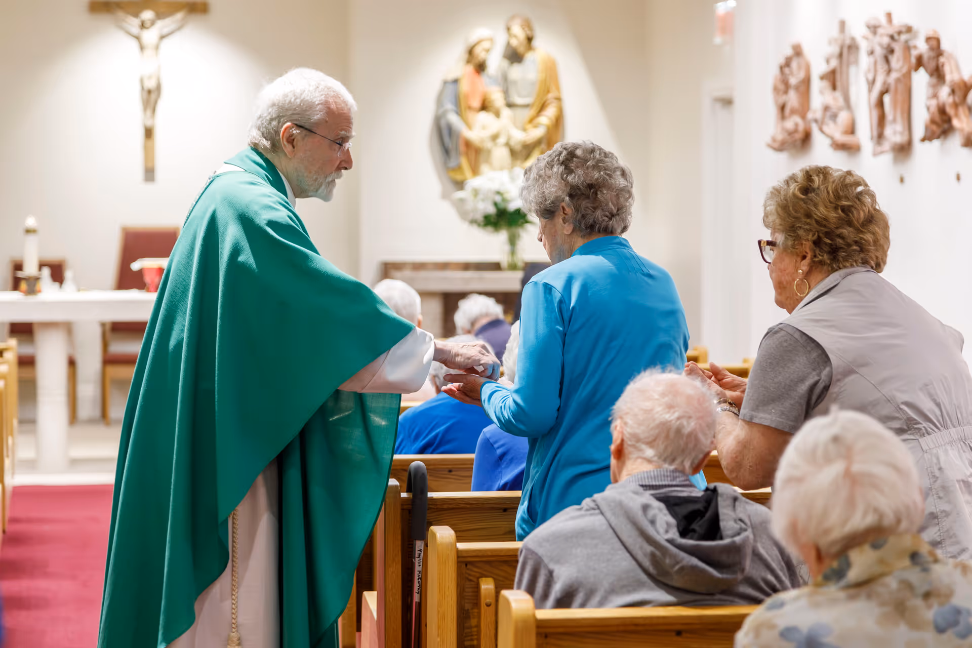 A priest in a green robe is giving communion to elderly people seated in wooden pews inside a chapel. Religious statues and a crucifix are visible on the walls in the background.