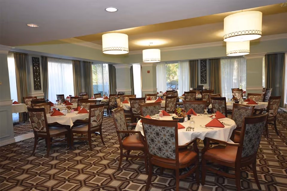 A dining room with multiple round tables covered with white tablecloths and set with red folded napkins, glasses, and silverware. The chairs have floral patterned upholstery. Large windows with curtains allow natural light to fill the room, and there are three large ceiling lights providing additional illumination. The floor is carpeted with a geometric pattern.
