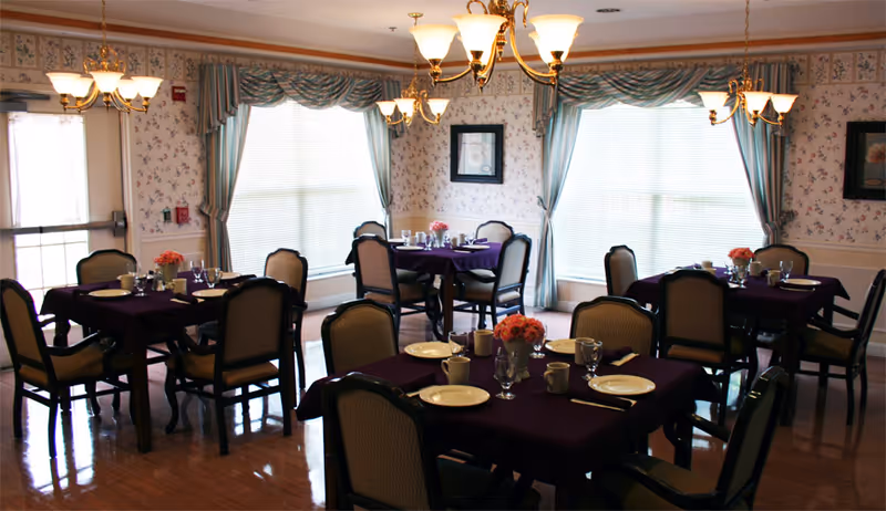 Dining room with multiple tables covered in dark purple tablecloths, each set with plates, cups, glasses, and silverware. The room has floral wallpaper, large windows with blue curtains, and chandeliers hanging from the ceiling providing warm lighting.