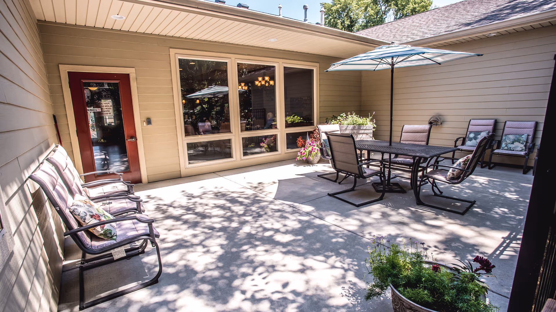 Enclosed sunny patio with a dining table, umbrella, multiple chairs, potted plants, and a red door leading into the building.
