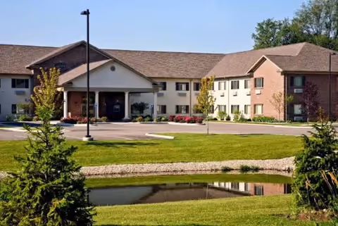 Exterior view of a two-story senior living facility building with a covered entrance, surrounded by a well-maintained lawn, small trees, and a pond in the foreground.