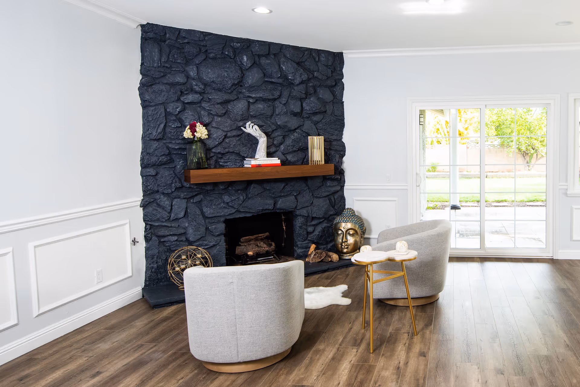 A modern living room with a dark stone fireplace wall featuring a wooden mantel decorated with a vase of flowers, a sculpture of a hand, and books. In front of the fireplace are two light gray armchairs and a small round side table with two candles. To the right, large sliding glass doors open to a green backyard.