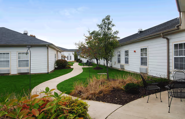 Outdoor courtyard area at Lorain Estates Senior Living with a winding concrete pathway, green grass, small trees, shrubs, and white single-story buildings on either side. There is a small patio area with black metal chairs and a table.