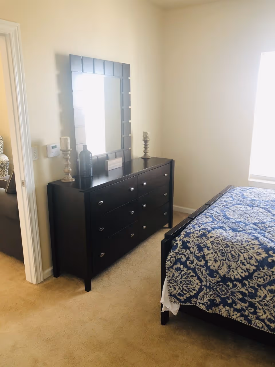 A bedroom with beige carpet featuring a dark wooden dresser with six drawers and a large rectangular mirror above it. On top of the dresser are two candle holders and a decorative bottle. To the right is a bed with a blue and white patterned bedspread. A window with bright light is visible on the right wall.