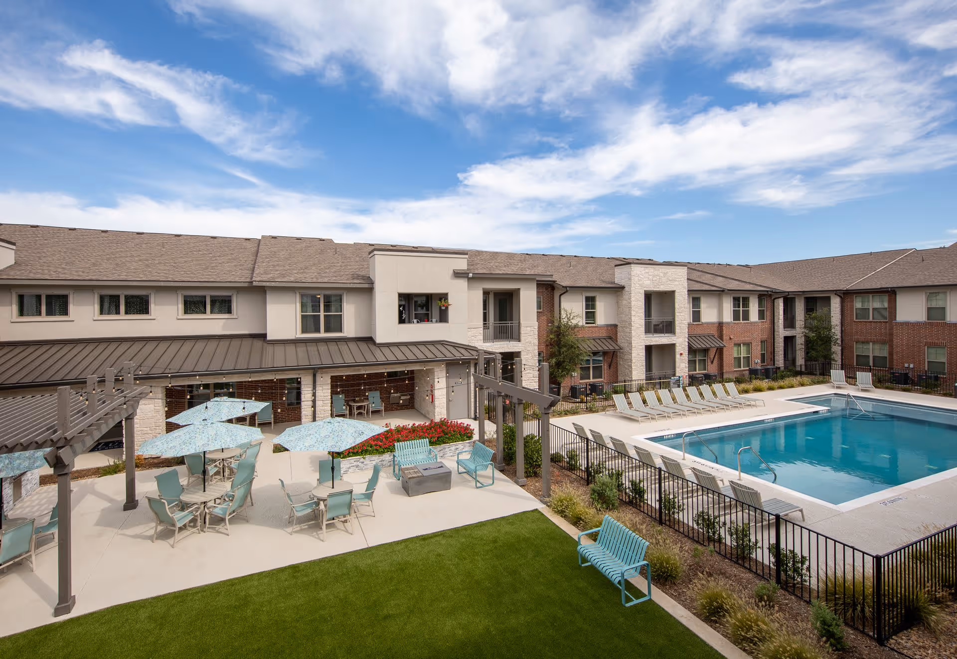 Courtyard with a swimming pool, lounge chairs, umbrellas and patio seating surrounded by two-story residential buildings under a blue sky.