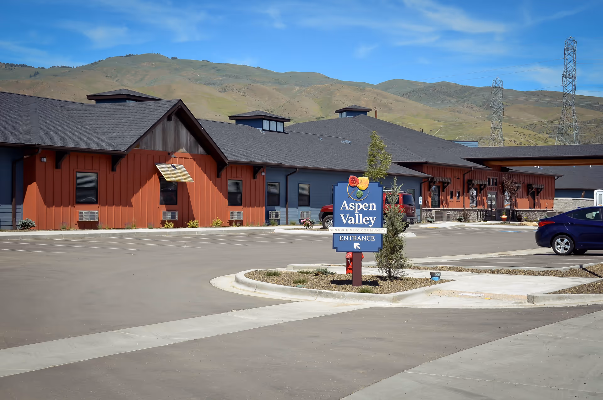 Exterior view of Aspen Valley Senior Living facility with a parking lot in front, a sign indicating the entrance, and hills in the background under a blue sky.