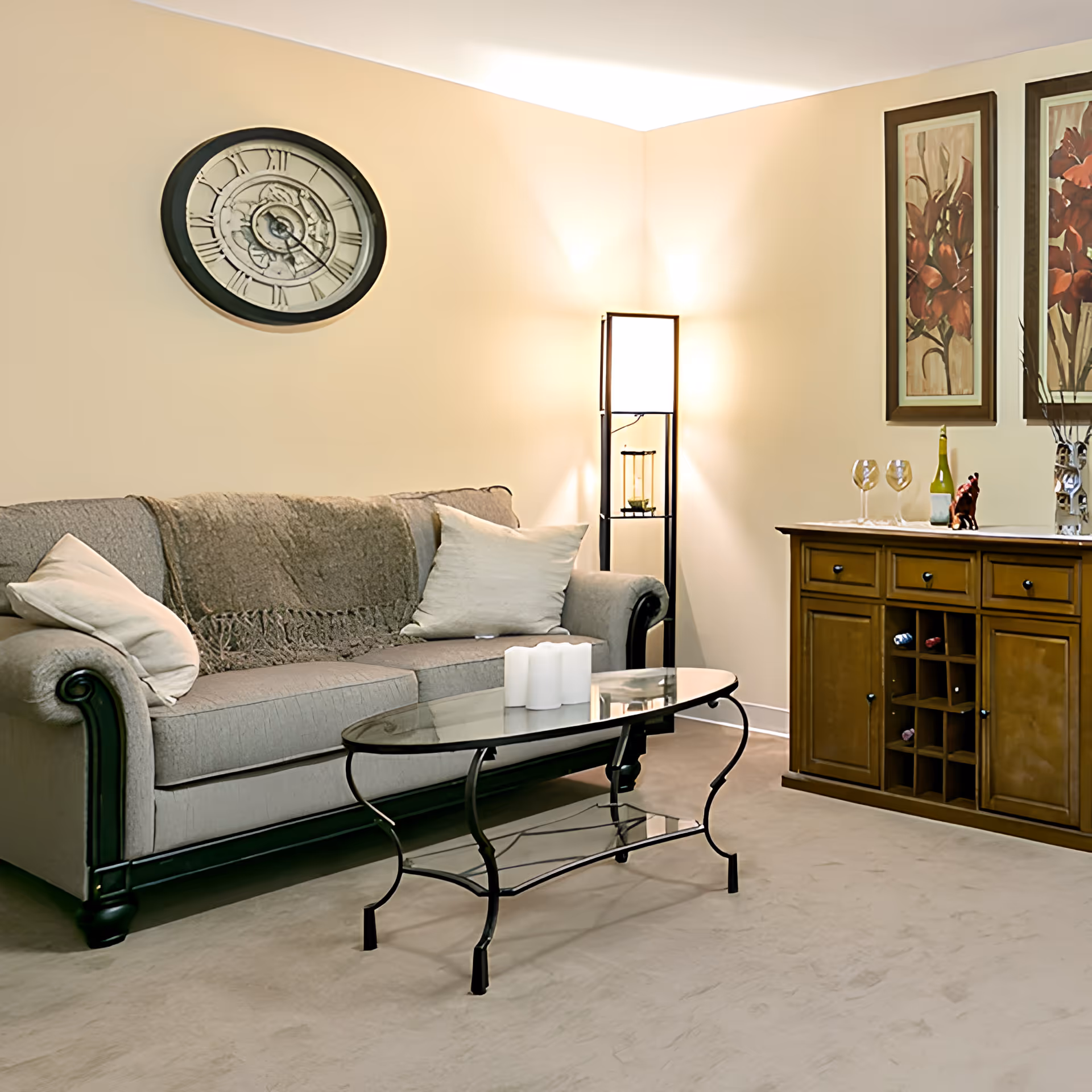 A cozy living room with a gray sofa adorned with two light-colored pillows and a throw blanket. A glass coffee table with three white candles sits in front of the sofa. A tall floor lamp is positioned in the corner, casting warm light. On the right side, there is a wooden cabinet with wine storage, topped with two wine glasses, a bottle of wine, and decorative items. Two framed floral paintings hang above the cabinet.