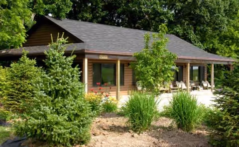Single-story building with front porch seating, surrounded by trees and landscaped plants.