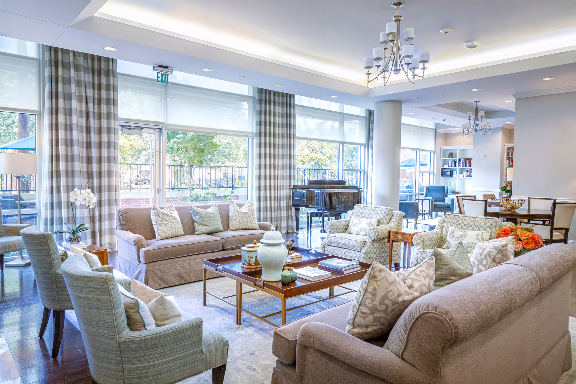 Bright and spacious living room area with large windows covered by checkered curtains, two beige sofas with patterned cushions, two light blue armchairs, a wooden coffee table with decorative items, a grand piano near the windows, and a dining area with chairs and a table in the background.