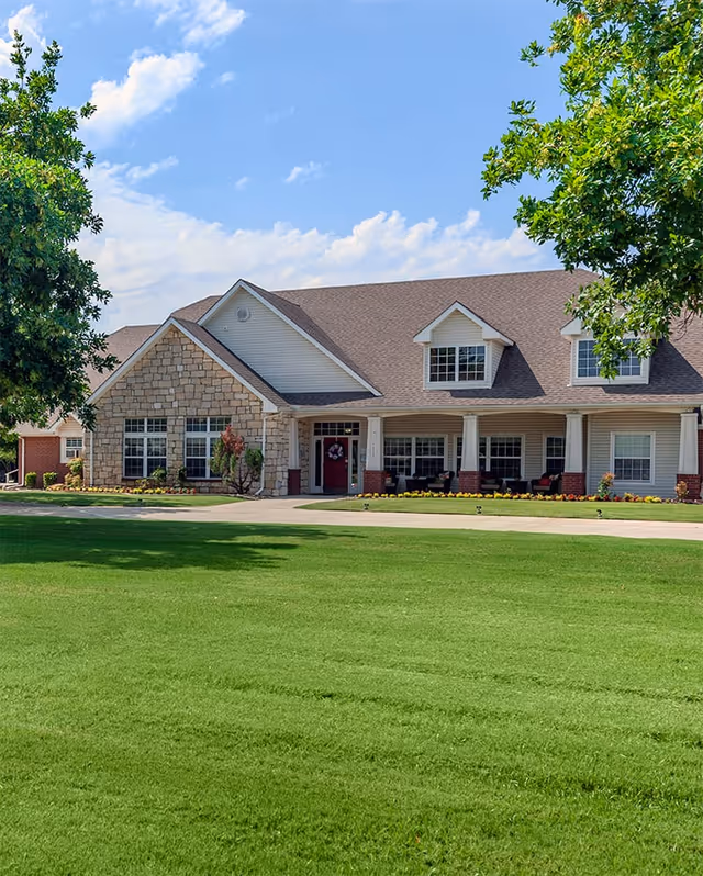 Front exterior of a single-story brick-and-siding senior living building with a covered porch, dormer windows, and a large green lawn.
