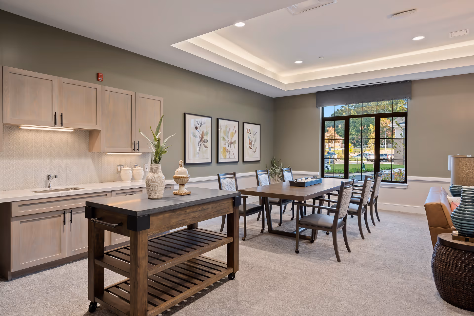 A well-lit interior room featuring a dining area with a wooden table and six chairs, a kitchen counter with light wood cabinets and a sink, a movable kitchen island with decorative vases, three framed botanical prints on the wall, a large window with a view of trees and parked cars outside, and part of a brown leather sofa with a side table and lamp.