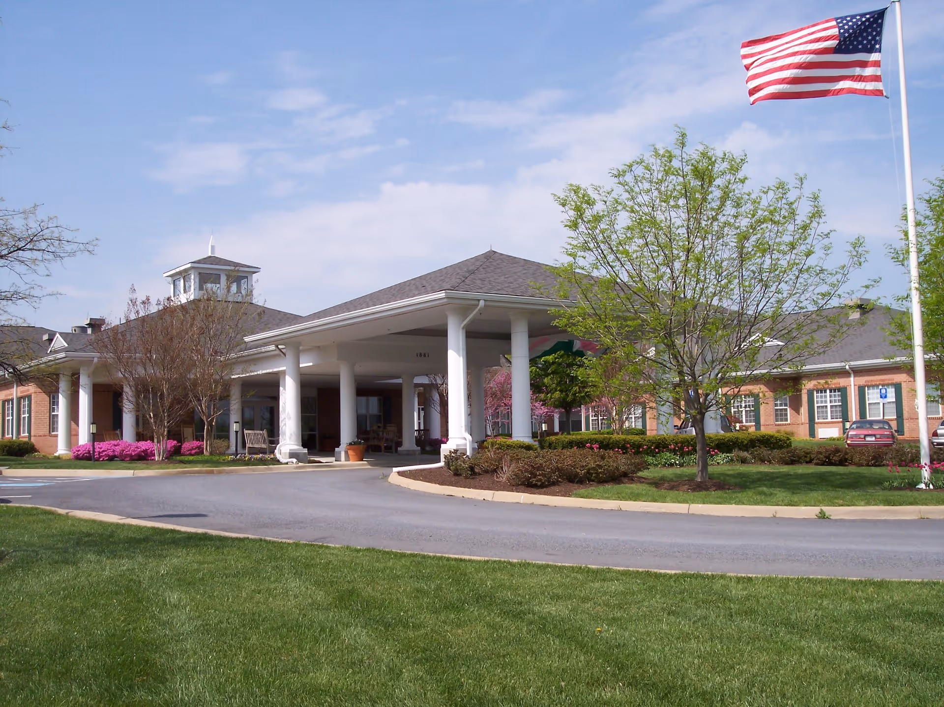Exterior view of The Willows At Meadow Branch facility showing a single-story brick building with a covered entrance supported by white columns. There is a circular driveway, well-maintained green lawn, trees, flowering bushes, and an American flag on a flagpole against a partly cloudy sky.