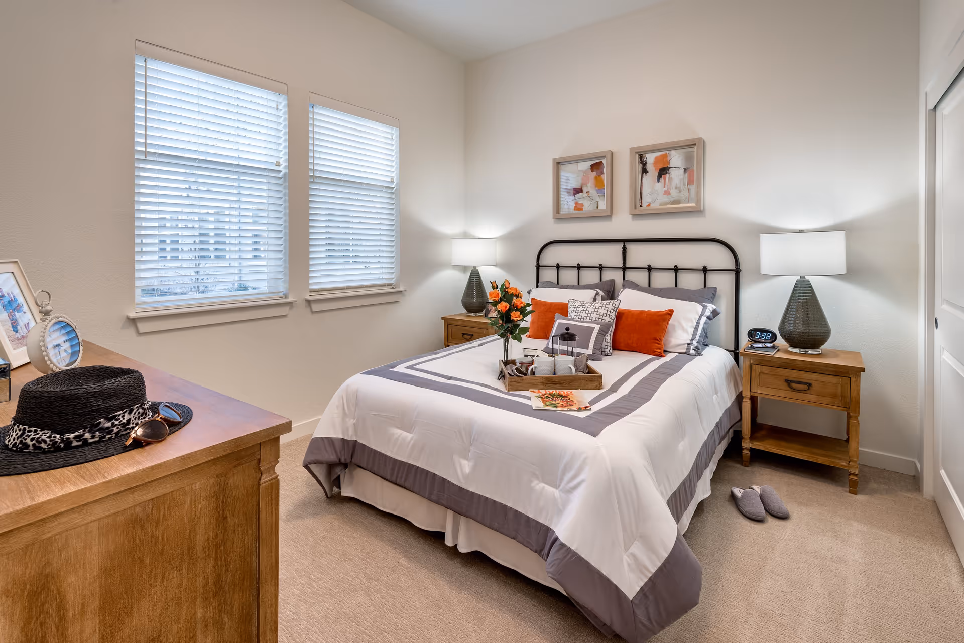 A cozy bedroom with a neatly made bed featuring white and gray bedding accented with orange pillows. There are two wooden nightstands on either side of the bed, each with a lamp. A tray with flowers, cups, and a lantern is placed on the bed. Two abstract paintings hang above the black metal headboard. A wooden dresser with a black hat and sunglasses is visible on the left side of the room. The room has beige carpet and two windows with white blinds.