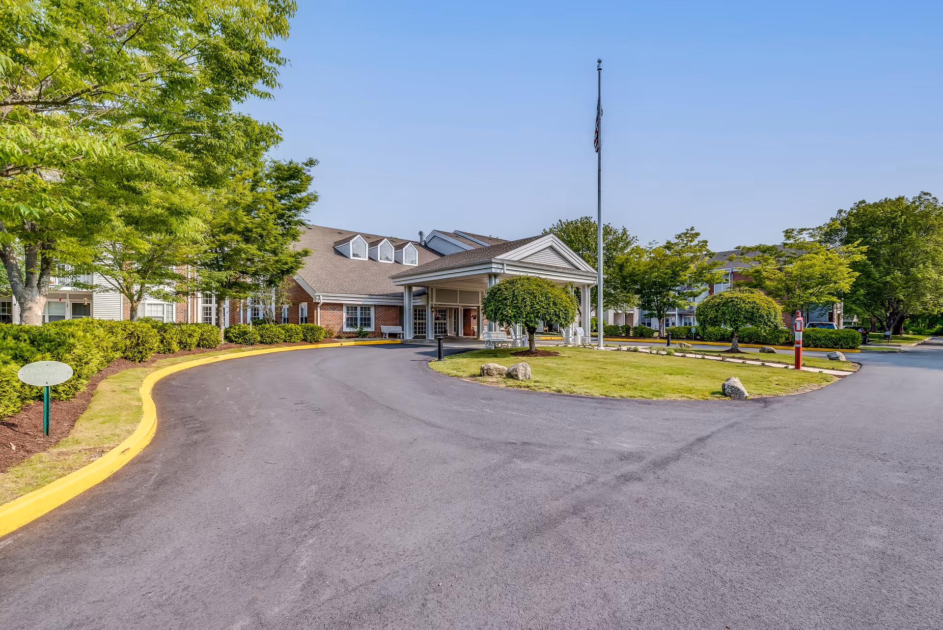 Exterior view of Solstice Senior Living at Groton showing a curved driveway leading to the main entrance with a covered portico. The building is surrounded by well-maintained greenery, trees, and bushes under a clear blue sky.