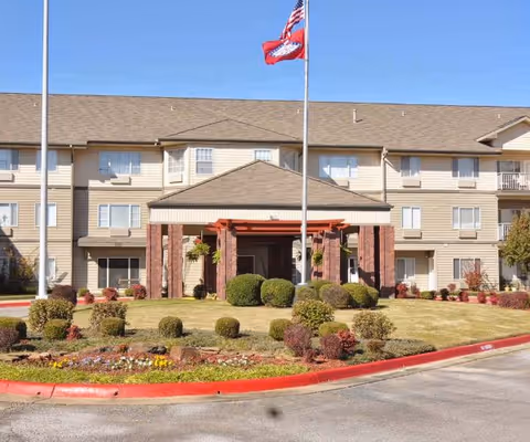 Front exterior view of a three-story senior living facility building with beige siding and brick accents. There is a covered entrance with columns, a landscaped area with bushes and flowers, and two flagpoles displaying the American flag and another flag. The sky is clear and blue.