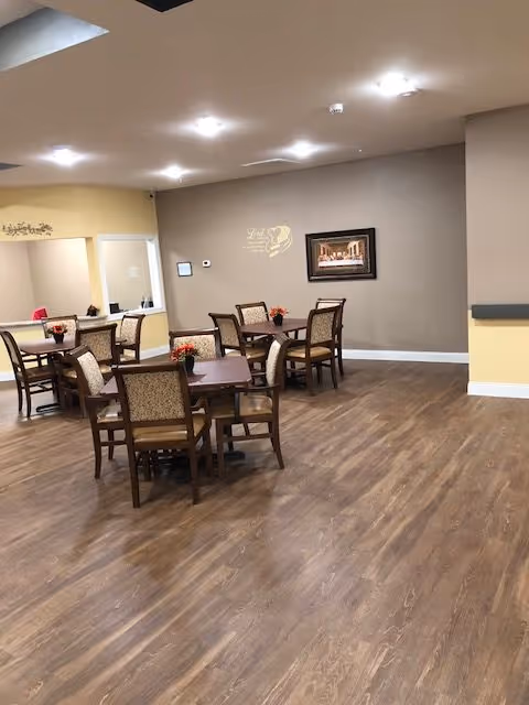 A dining area in a retirement facility with several wooden tables and chairs arranged on a wood-patterned floor. Each table has a small floral centerpiece. The walls are painted in neutral tones with a framed picture and a decorative wall decal. The ceiling has multiple recessed lights providing bright illumination.