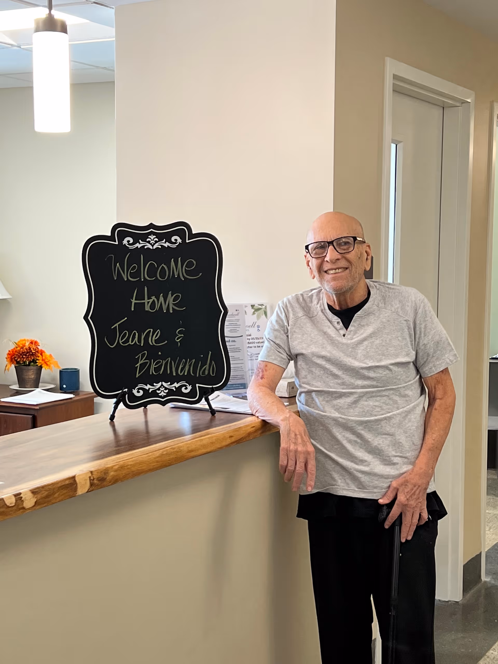 An elderly man with glasses and a cane is standing and smiling next to a wooden counter inside a facility. On the counter, there is a decorative blackboard sign that reads 'Welcome Home Jeane & Bienvenido'. The background shows a light-colored wall, a door, and some papers and a small flower pot on a side table.