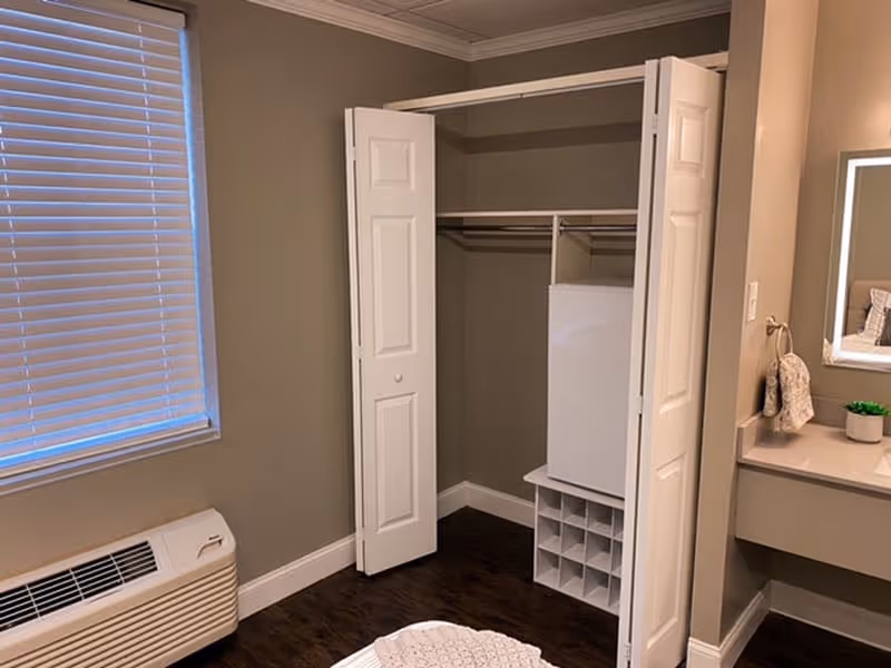 A corner of a senior living bedroom featuring an open white closet with hanging rods and shelves, a window with closed blinds, a heating/cooling unit below the window, and a vanity area with a mirror and small plant on the right side.