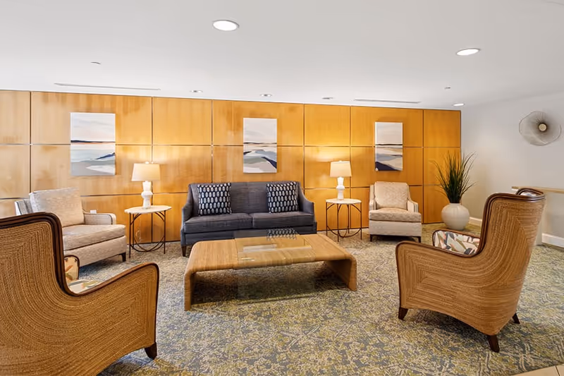 A cozy living room with a blue sofa, two beige armchairs, and two wicker chairs arranged around a wooden coffee table. The room features a patterned carpet, wooden paneling on the wall with three abstract paintings, two side tables with lamps, and a large potted plant in the corner.